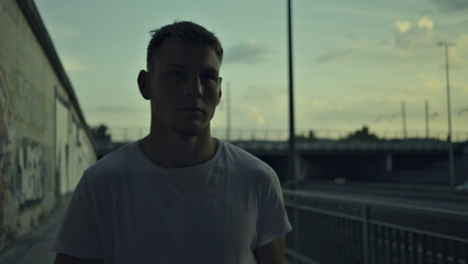 Contré-jour lighting highlights a young man's resolute stance in an urban setting, surrounded by graffiti and an overpass. His focused expression conveys resilience and inner strength.