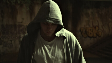 Close-up of a young man in a hooded sweatshirt, standing contemplatively in a graffiti-covered, dim alley, symbolizing urban resilience and introspection.