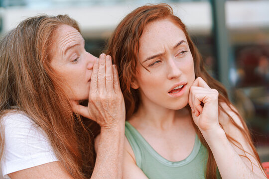 Two women mother and daughter engage in close conversation with one whispering while the other looks skeptical, capturing a moment of intrigue and curiosity in an outdoor setting