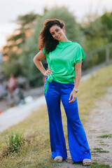 A cheerful woman in a bright green top poses with a warm smile, set against a backdrop of greenery, showcasing vibrant style and joyful spirit.