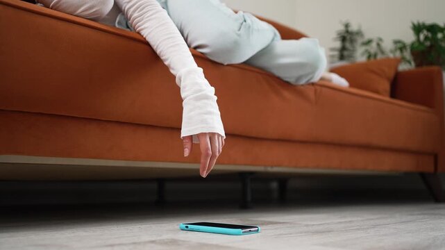 Mobile phone dropping from drowsy female's grasp, falling onto hardwood floor while reclining lazily on cushioned living room couch during quiet afternoon