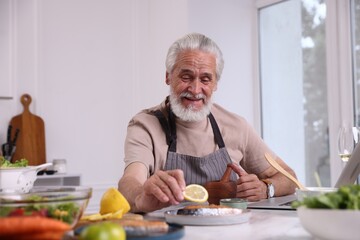 Elderly man putting slice of lemon onto salmon fish at white table indoors