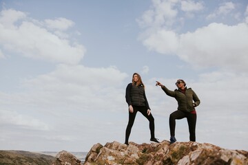 Two people stand on a rocky hill under a cloudy sky, wearing outdoor jackets. One points into the...