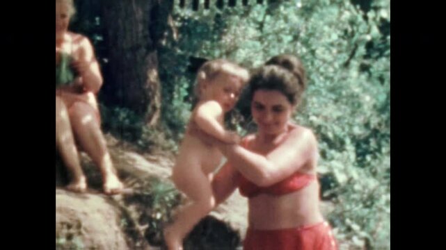 Grandmother gently hands baby to mother on sandy summer beach near river. Touching multi generational countryside vacation. Nostalgic family outdoors. Vintage film. Old archive. Archival retro 1960s