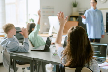 Group of teenagers sitting at desks raising hands while using laptops in classroom, female teacher standing in front of students engaging with class activity