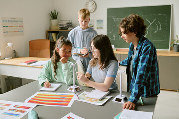 Group of diverse teenagers collaborating on science project in classroom, Caucasian girl explaining wind turbine model while Asian girl, Caucasian boy and another teenager listening and taking notes