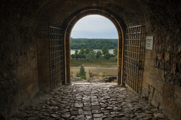 Ancient stone gateway opens to a scenic river landscape with greenery under a cloudy sky, framed by rustic wooden doors.