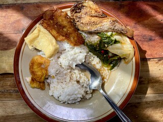 Heavy foods such as white rice with shrimp, salted fish, tofu, tempeh and sour vegetables are served on a ceramic plate on a wooden table, the eating process is almost finished, Indonesian staple food