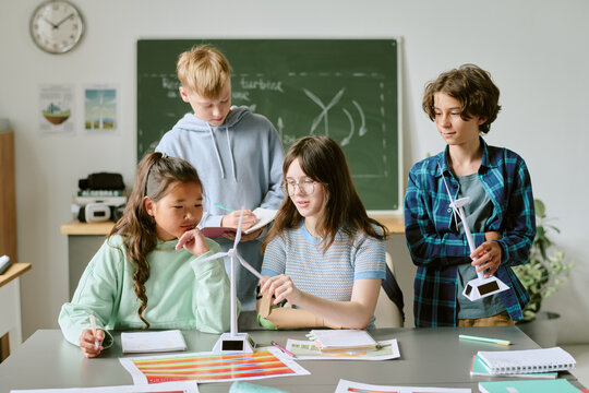 Group of multiethnic teenagers collaborating on science project, assembling wind turbine models and discussing renewable energy concepts in classroom with educational materials visible