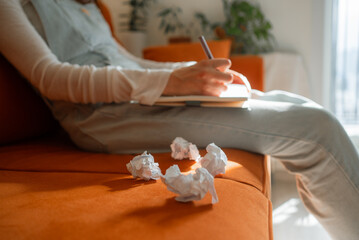 Female writer drafting notes on notebook while resting on orange sofa, discarded paper drafts surrounding workspace, illustrating creative brainstorming