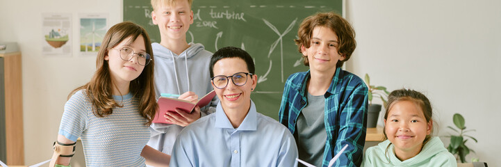 Website banner with portrait of group of diverse teenagers and Caucasian middle aged teacher smiling and posing together in classroom, teenagers holding notebooks and wind turbine models