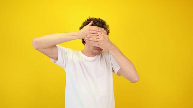 Shocked man covers his face with hands, peering fearfully through fingers against a yellow studio backdrop - perfect for Halloween promotions, horror films, and mental health awareness.