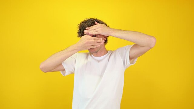 Shocked man covers his face with hands, peering fearfully through fingers against a yellow studio backdrop - perfect for Halloween promotions, horror films, and mental health awareness.