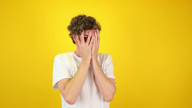 Shocked man covers his face with hands, peering fearfully through fingers against a yellow studio backdrop - perfect for Halloween promotions, horror films, and mental health awareness.