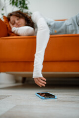 Young woman, possibly a student or freelancer, lies asleep on an orange sofa, her arm dangling towards a smartphone that has fallen onto the floor