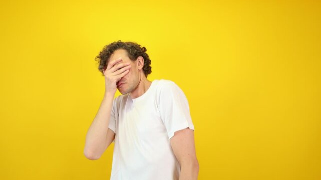 Shocked man covers his face with hands, peering fearfully through fingers against a yellow studio backdrop - perfect for Halloween promotions, horror films, and mental health awareness.