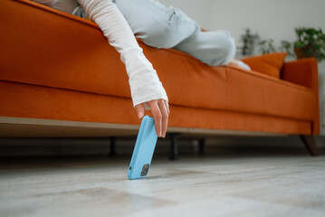 Woman lying on an orange sofa trying to reach her blue smartphone that has fallen on the floor, depicting a common scenario of accidental phone drops at home