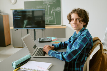 Portrait of Caucasian teenage boy sitting at desk using laptop and looking at camera in classroom setting with notebook and pen case on table, digital screen in background