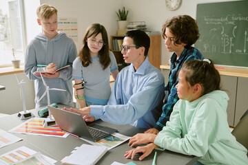 Caucasian male teacher explaining project to group of teenagers, students listening and collaborating around laptop in classroom