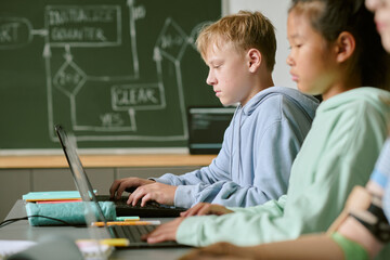 Caucasian boy and Asian girl teenagers sitting side by side using laptops in classroom, focusing on typing and working on assignments with another teenager partially visible in foreground