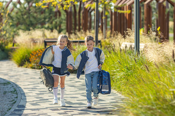 Sibling, happy little boy and girl, both schoolchildren, run joyfully through a city park after school lessons, each carrying their backpacks. Friendship and back to school concept.