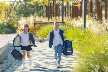 Sibling, happy little boy and girl, both schoolchildren, run joyfully through a city park after school lessons, each carrying their backpacks. Friendship and back to school concept.