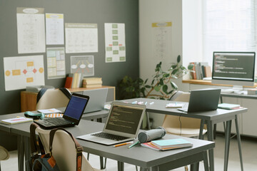 Empty classroom featuring several laptops displaying code on desks, notebooks and stationery scattered around, sunlight streaming through window, educational posters on wall