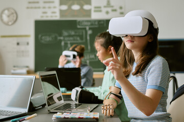 Caucasian teenage girl wearing virtual reality headset interacting with digital interface in classroom, Asian teenage girl using laptop and VR headset in background, technology education concept