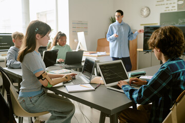 Group of multiethnic teenagers sitting at desks using laptops and writing in notebooks while...