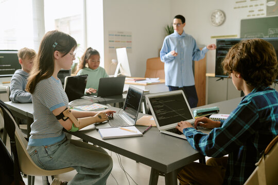 Group of multiethnic teenagers sitting at desks using laptops and writing in notebooks while teacher standing near chalkboard explaining lesson in classroom setting