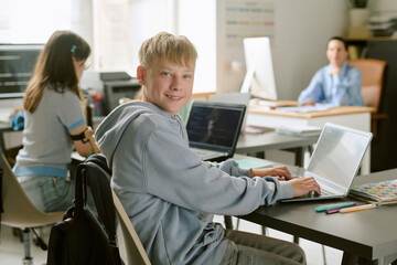 Portrait of Caucasian teenage boy smiling while using laptop at desk in classroom, other teenagers studying in background, modern educational environment with technology focus