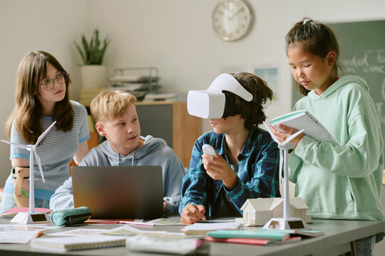 Group of multiethnic teenagers collaborating on science project, one Caucasian boy wearing virtual reality headset while others observing and taking notes, wind turbine models on table