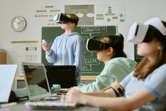 Group of Caucasian teenagers using virtual reality headsets and controllers in classroom, sitting and standing at desks with laptops, engaging in interactive educational activity together