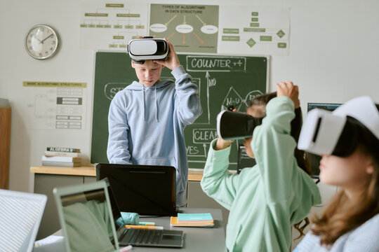 Teenage Caucasian boy standing using laptop while adjusting virtual reality headset, two teenage girls wearing VR headsets sitting at desk in classroom with educational posters and chalkboard