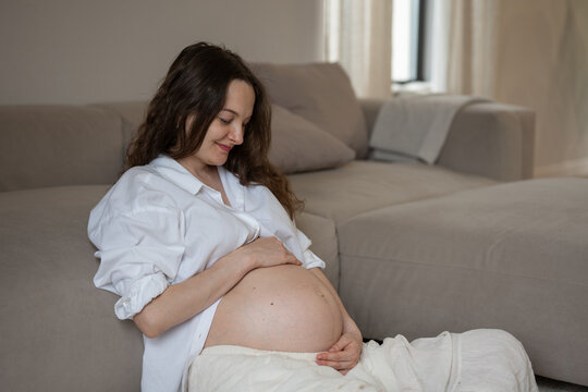 Smiling pregnant woman caressing her belly while sitting on the floor near the sofa in the living room at home