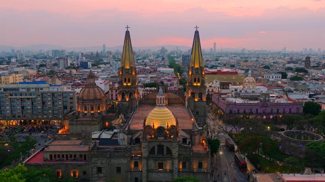 Aerial dolly in flight toward illuminated dome and spires of Guadalajara Cathedral at sunset, flight reveals crowded Plaza de Armas below