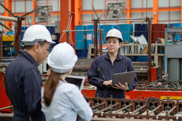 Three workers in a factory discuss a project using laptop computer. Everyone wearing protective hardhat. Atmosphere in manufactory with large machinery.