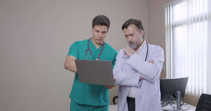 Young male medical staff presenting information on laptop to senior caucasian doctor during teamwork session inside hospital office showing collaboration and clinical decision-making process
