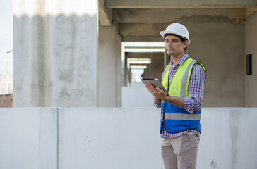 Young caucasian worker in hardhat and safety vest review plan on tablet computer. Working atmosphere at a construction site with unfinished concrete wall.