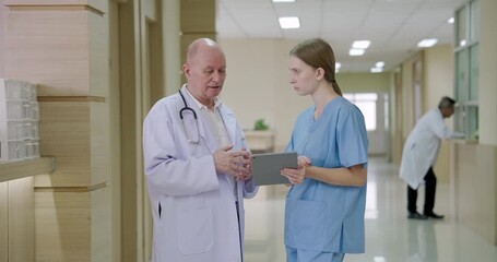 senior caucasian male doctor discussing medical case with young caucasian female nurse using digital tablet standing near nurse station inside hospital hallway during healthcare team coordination - Powered by Adobe