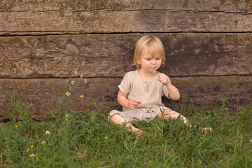 A happy blond boy with long hair is sitting on the grass by a wooden wall with a flower in his hands