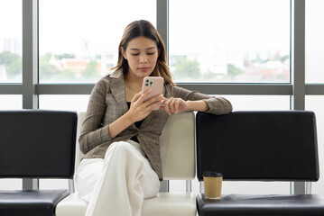 A woman dressed in professional clothing sit in an airport lounge, focused on her smartphone. She has a coffee cup beside her and appear relaxed while waiting for her flight.