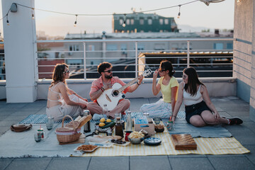 Diverse friends gathers on a rooftop to enjoy a relaxing picnic, playing music, chatting, and enjoying snacks. The setting is adorned with string lights, creating a cozy atmosphere at sunset.