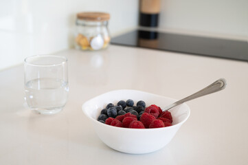 A bowl of mixed berries and a glass of water sit on a counter. The bowl is filled with blueberries and raspberries, and the glass is half full of water. Concept of health and wellness