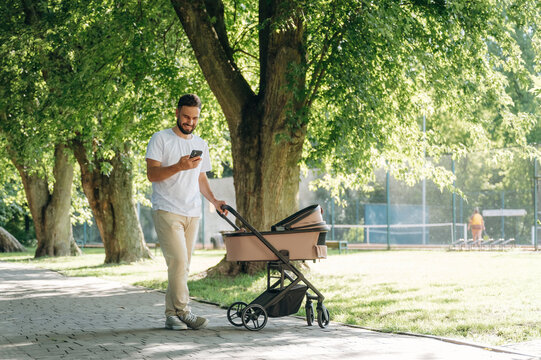 Using smartphone. Father is with cradle, having a walk in the park - Powered by Adobe