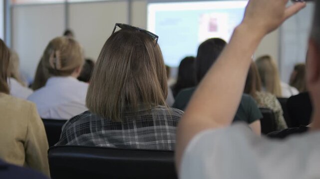 Group of people are sitting in a classroom. A woman is sitting in the front row. A man is sitting in the back row