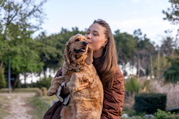 A young Caucasian woman with long brown hair kisses a golden cocker spaniel. They are outdoors in a park with greenery in the background.