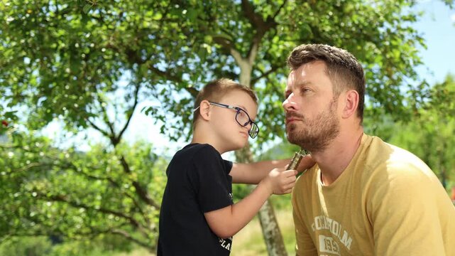 Sweet boy with down syndrome trimming his father's beard outside in the yard. summer, family, vacation, nature, real authentic people. Fun, emotions, love. Candid diverse people. 