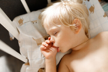 small cute blonde child sleeping in his white crib and sucking his thumb finger. sucking reflex