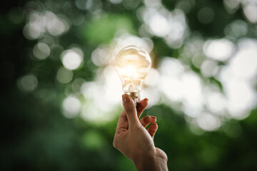 A hand holds a glowing light bulb against a green, blurred background, symbolizing innovation, creativity, eco-friendly ideas, energy, inspiration, and sustainable thinking in nature.